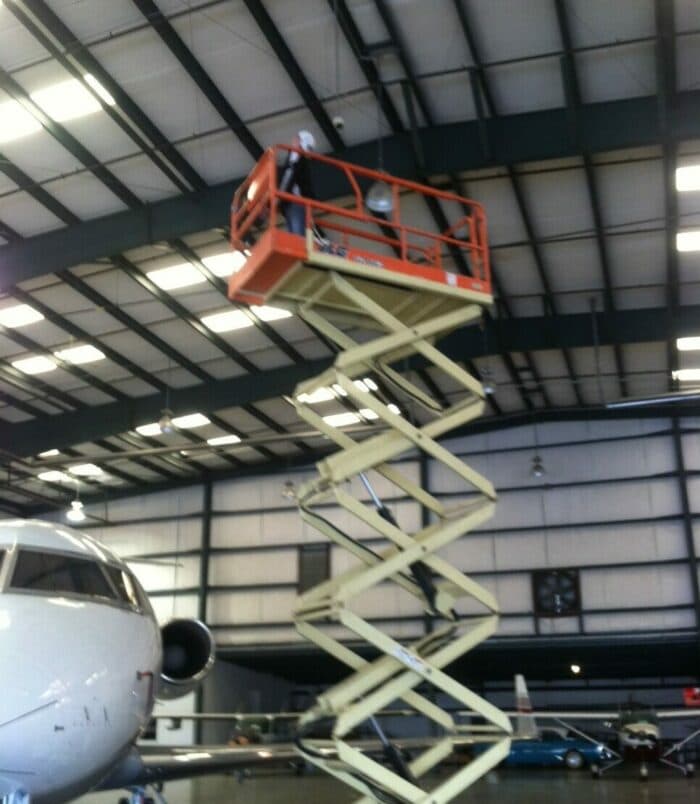 A scissor lift extended to the roof of a hanger. A man is working on the roof next to a private jet.